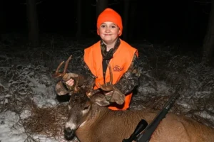 A young boy in an orange vest is holding his deer.