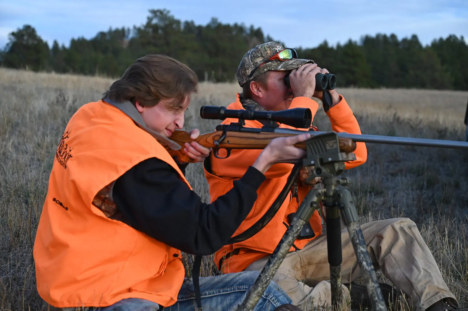 Two people in orange vests looking through a scope