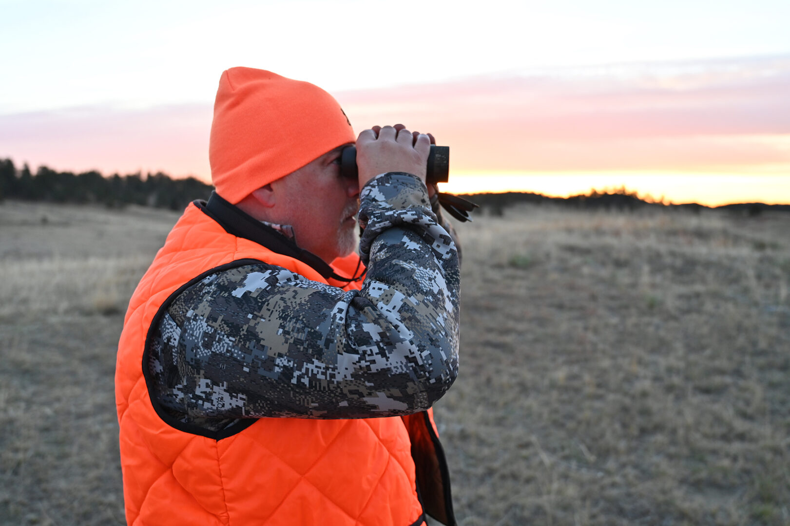 A man in an orange vest looking through binoculars.