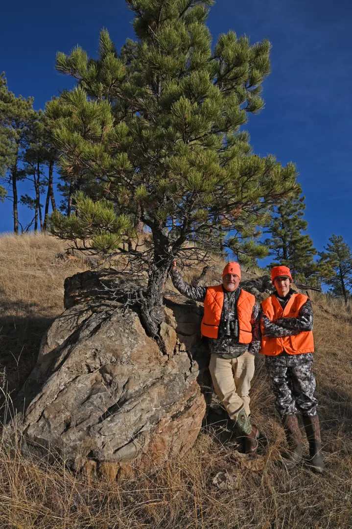 Two men in orange vests standing next to a tree.