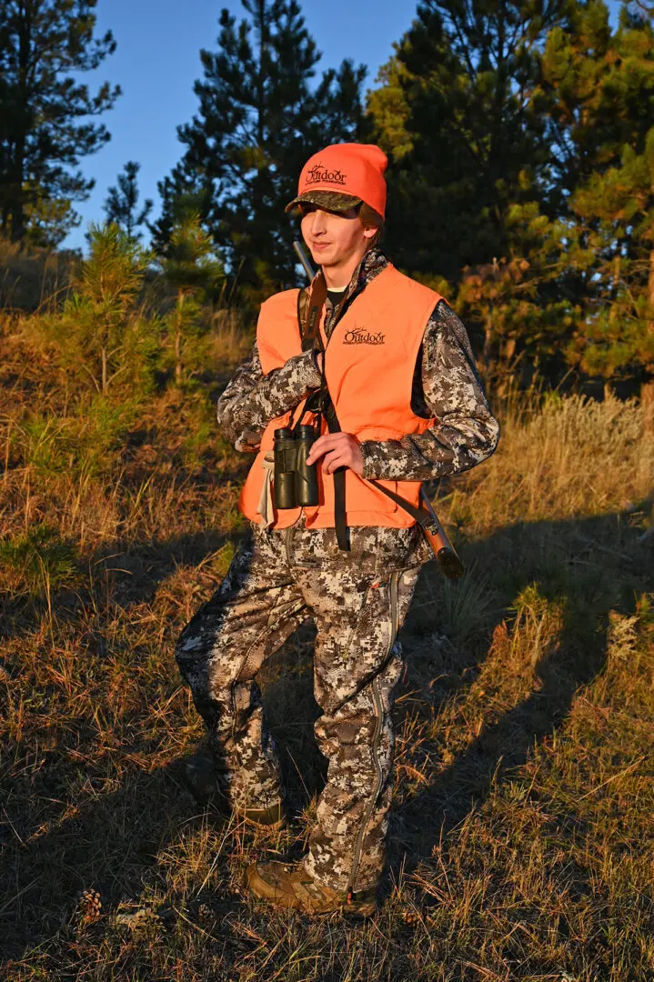 A man in camouflage and an orange vest holding a camera.