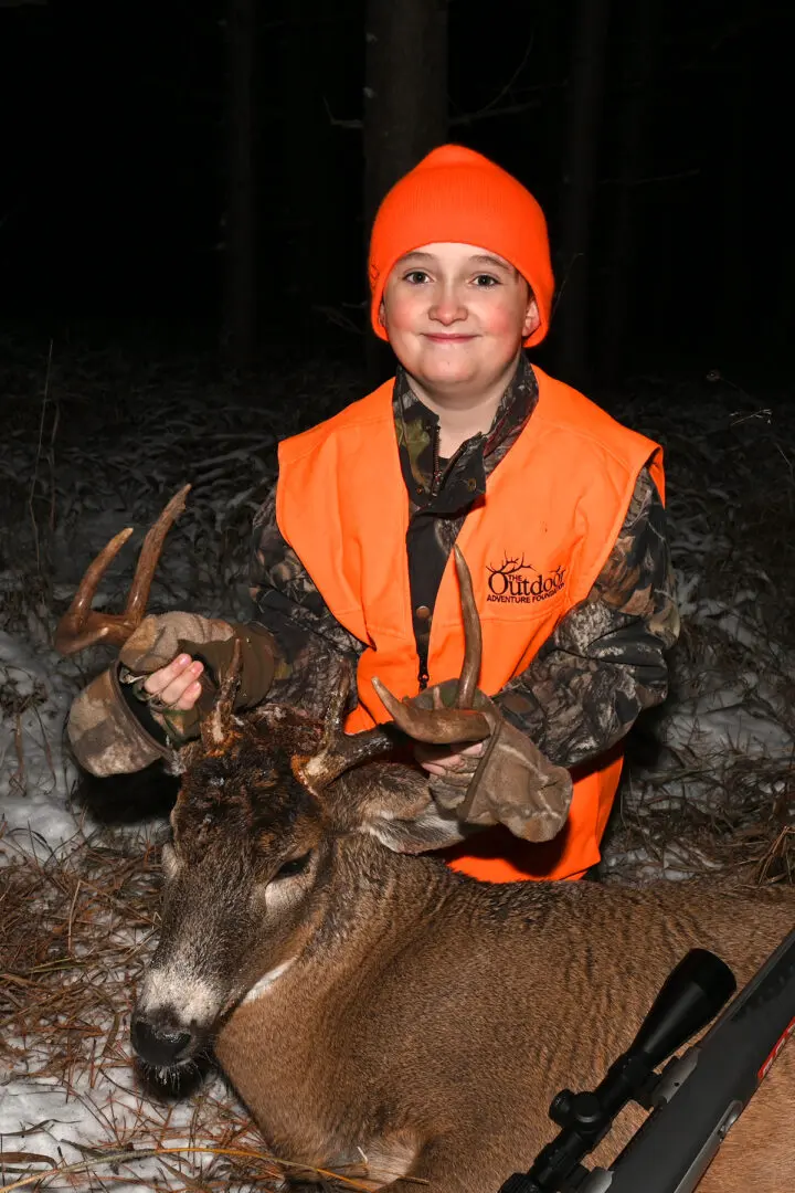 A young boy in an orange vest holding two deer.