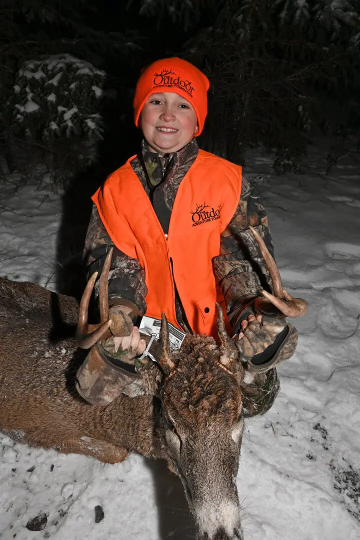 A young boy in an orange vest is holding two deer.
