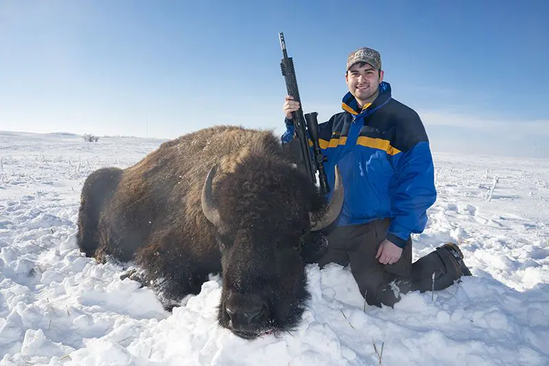 A man holding an automatic rifle next to a bison.