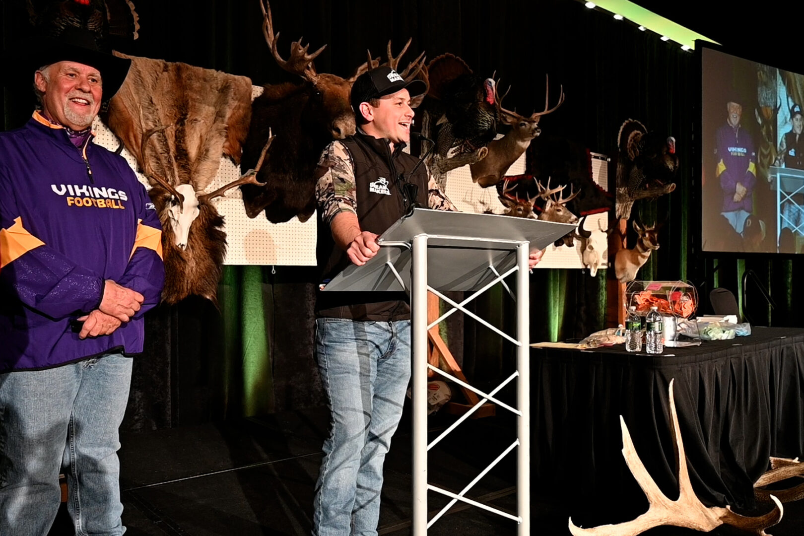 A man standing at a podium with many deer on the wall behind him.