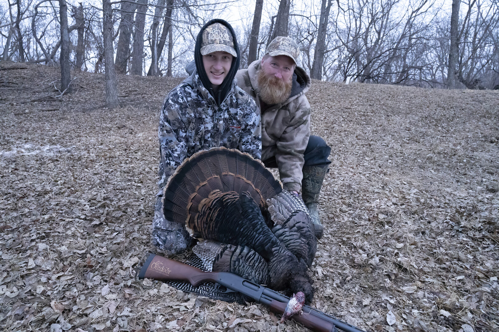 Two people posing with a turkey in the woods.