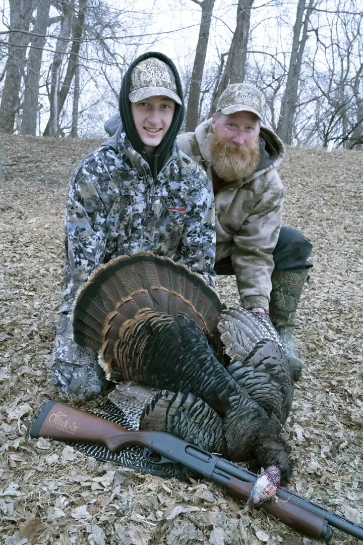 Two men pose with a turkey in the woods.