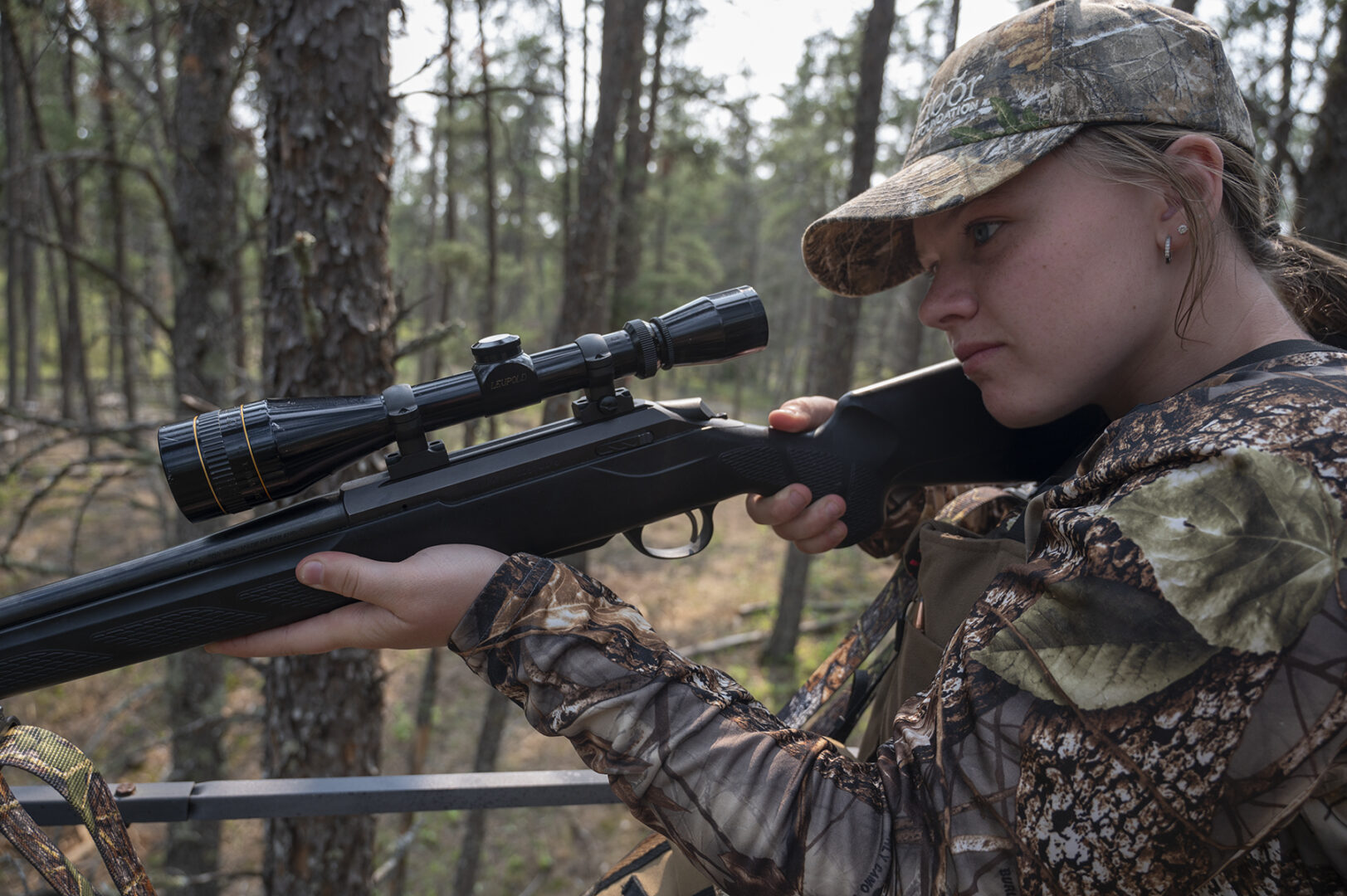 A woman in camouflage holding a rifle and looking through the scope.