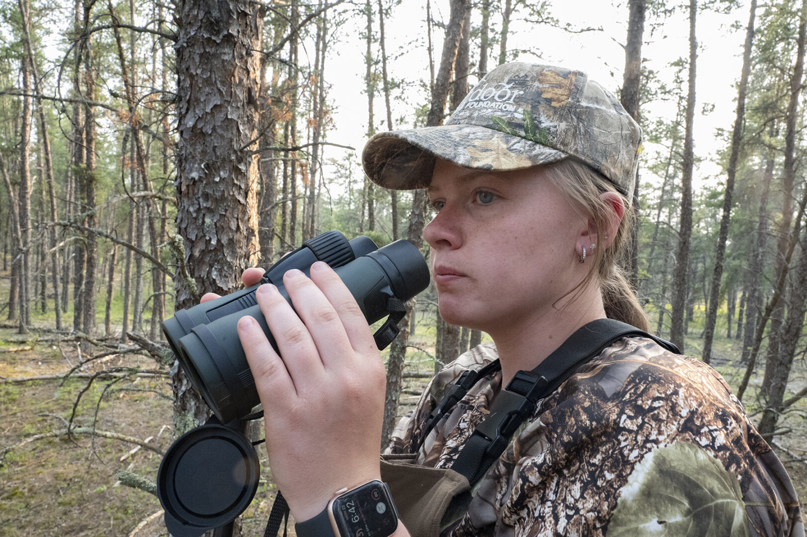 A woman in camouflage holding binoculars while standing next to trees.