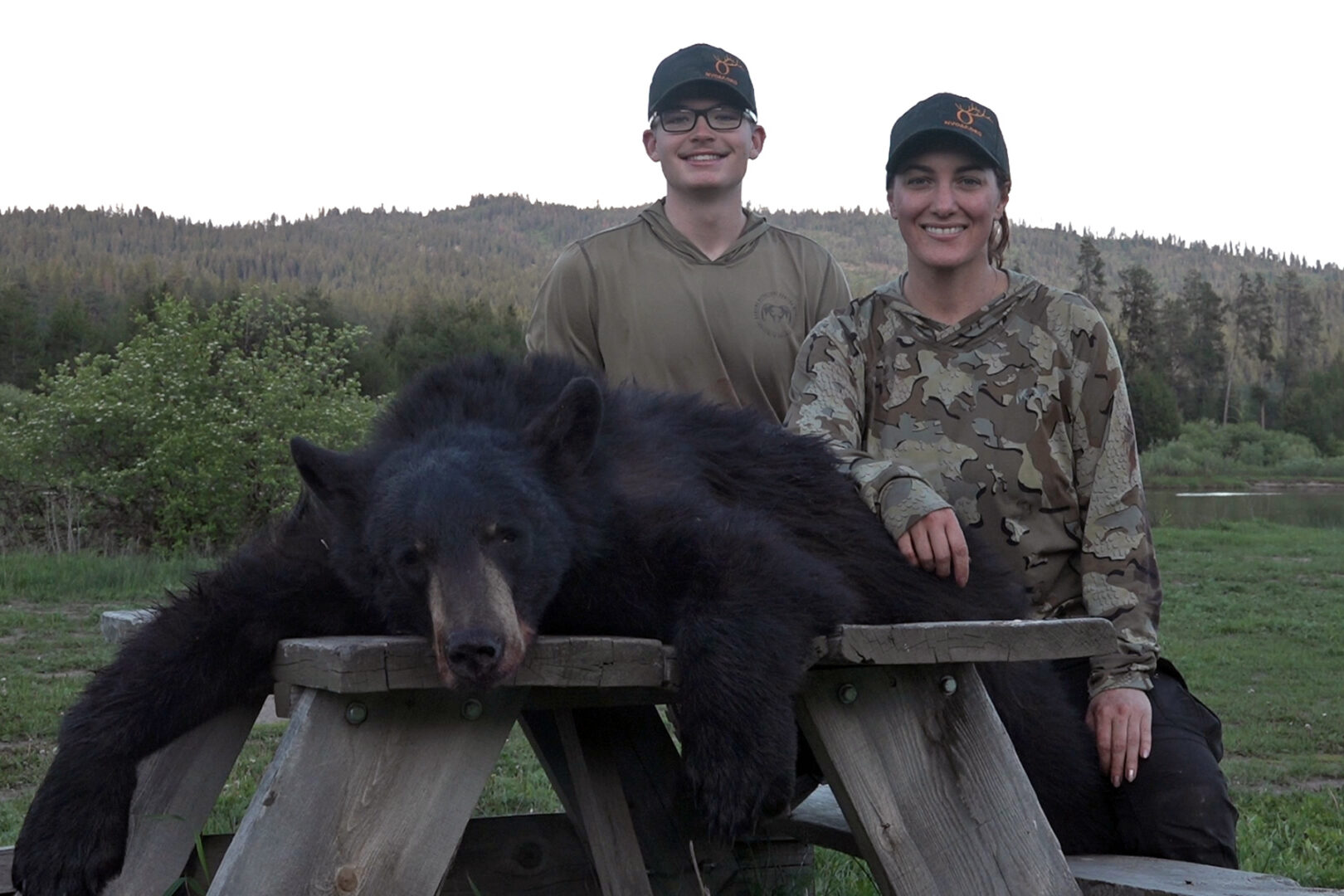 Two people posing with a black bear on top of a wooden fence.