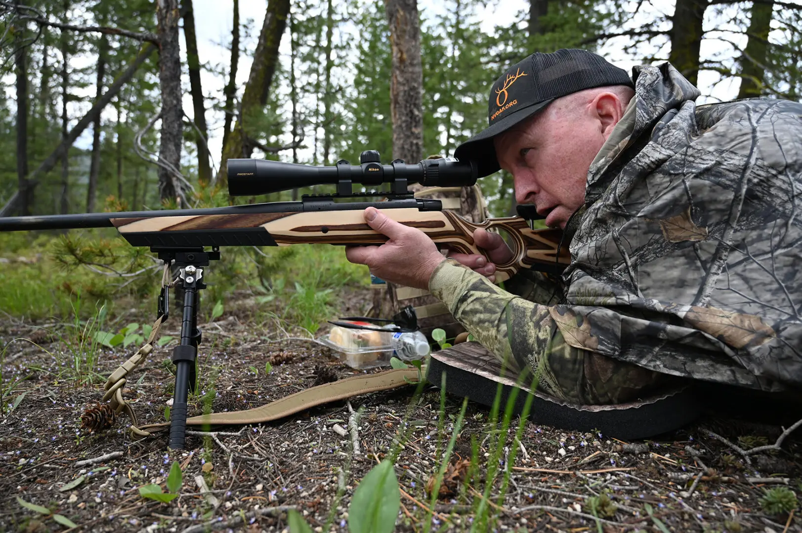 A man in camouflage laying on the ground aiming with a rifle.