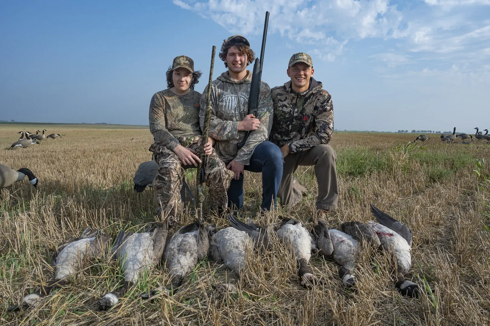Three hunters pose with their guns while a group of ducks lay in the grass.