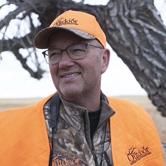 A man in an orange jacket and hat standing next to a tree.